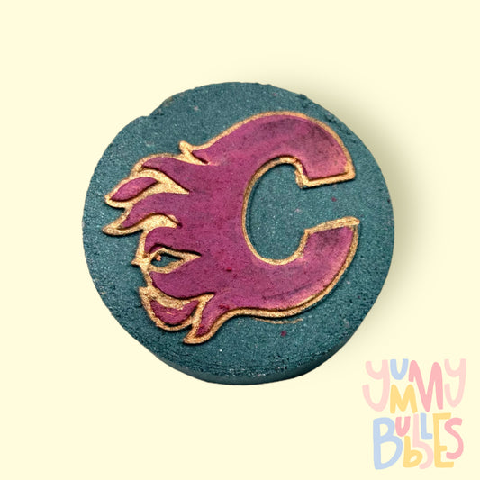 Hockey puck shaped bath bomb with a Calgary Flames logo in the center. Painted with red and gold and is a black bath bomb
