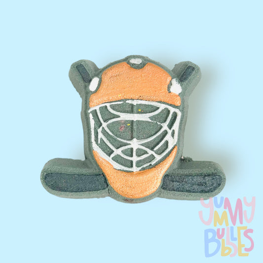 Black hockey helmet shaped bath bomb with hockey sticks in the background. The helmet is painted orange and white and the hockey stick is painted black.