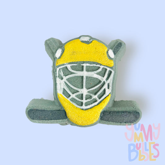 Black hockey helmet shaped bath bomb with hockey sticks in the background. The helmet is painted yellow and white and the hockey stick is painted black.
