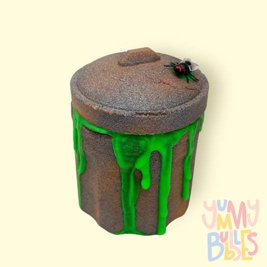 Grey trashcan shaped bath bomb highlighted with a brownish color with green ooze coming out from the lid and a toy fly on top